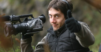 A photograph of David Proud holding a camera and making a thumbs up sign.  He is in an Outdoor location.