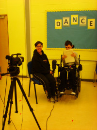 Image shows Hillie being filmed interviewing one of her classmates. You can see the video in the forground. Hillie is holding a large fluffy microphone 