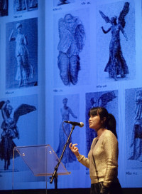 Jewlry designer Lin Cheung stands behind a transparent lecturn, in a gold top, in front of a blue backdrop which features pictures of the goddess of victory, Nike.